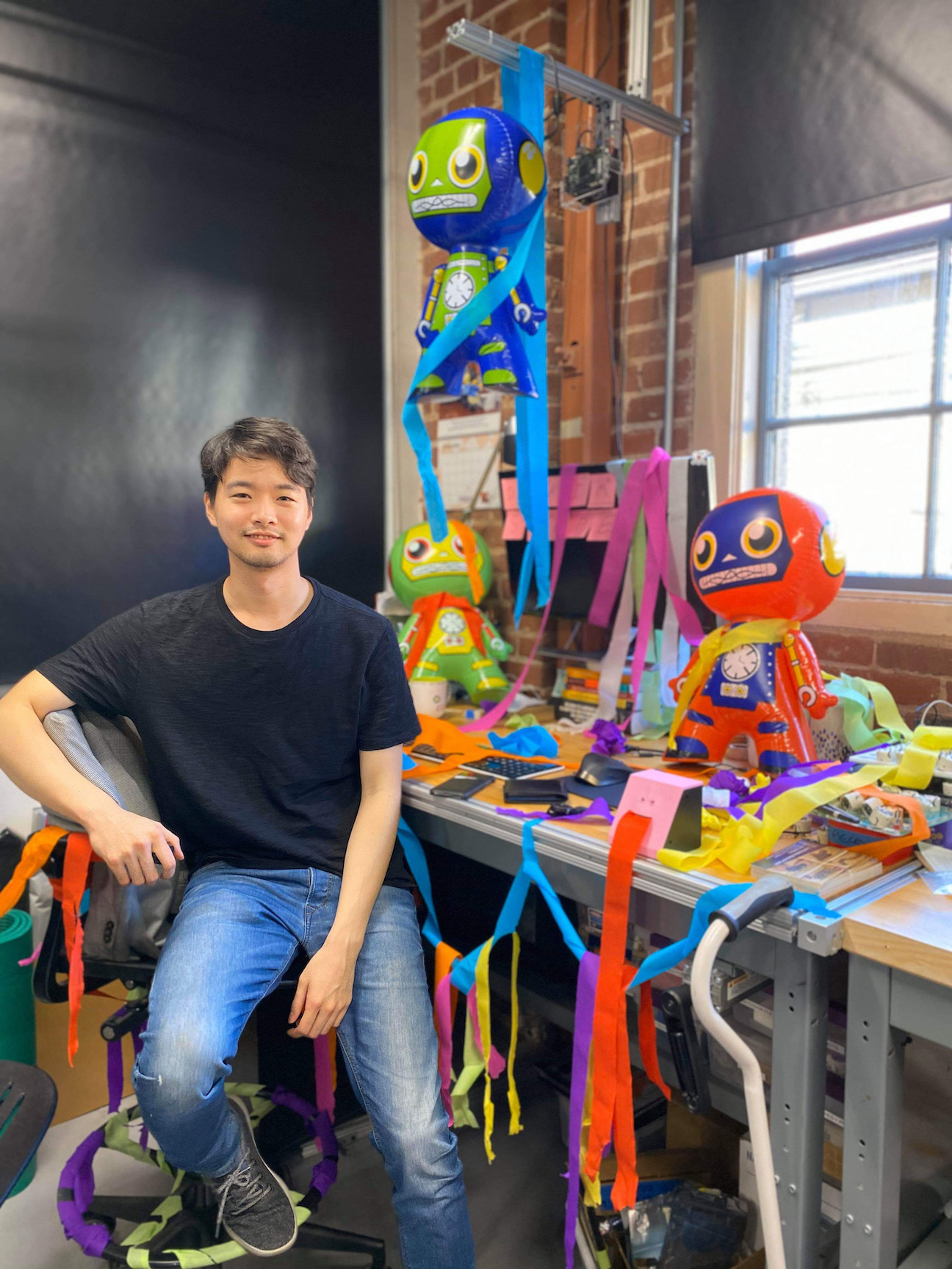 picture of Lawrence standing in front of his desk full of colorful papers and inflatable robots.